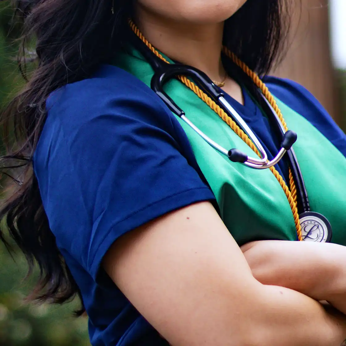 A person wearing a blue outfit and a green sash holds a stethoscope around their neck with arms crossed. The image conveys professionalism and confidence.