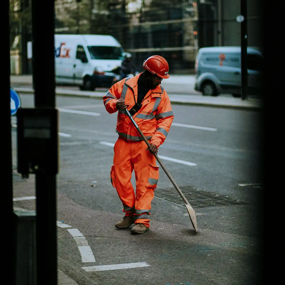 A construction worker in bright orange gear and a helmet uses a spade on an urban road. Vans are parked nearby, suggesting an active city setting.