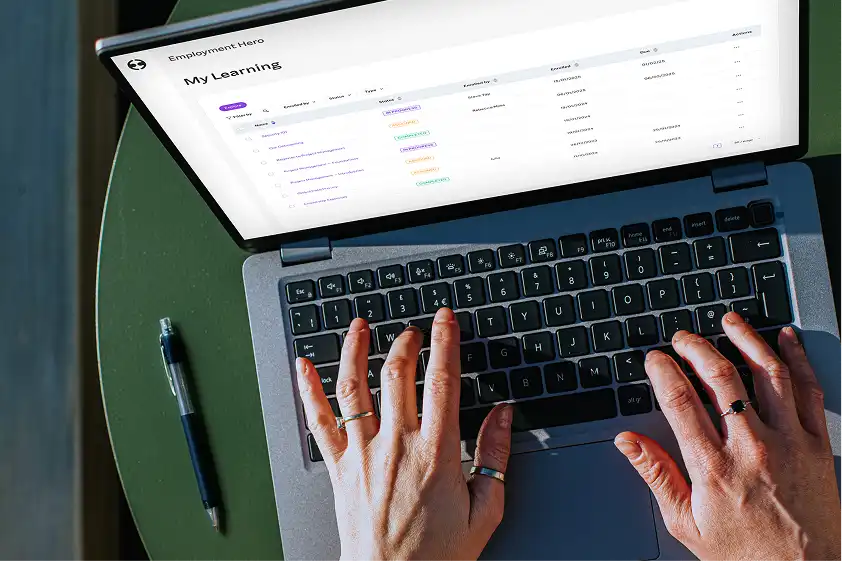 Hands typing on a laptop displaying a "My Learning" dashboard. The screen shows a list of courses with colorful status labels. A pen lies beside the laptop, suggesting a work or study environment.